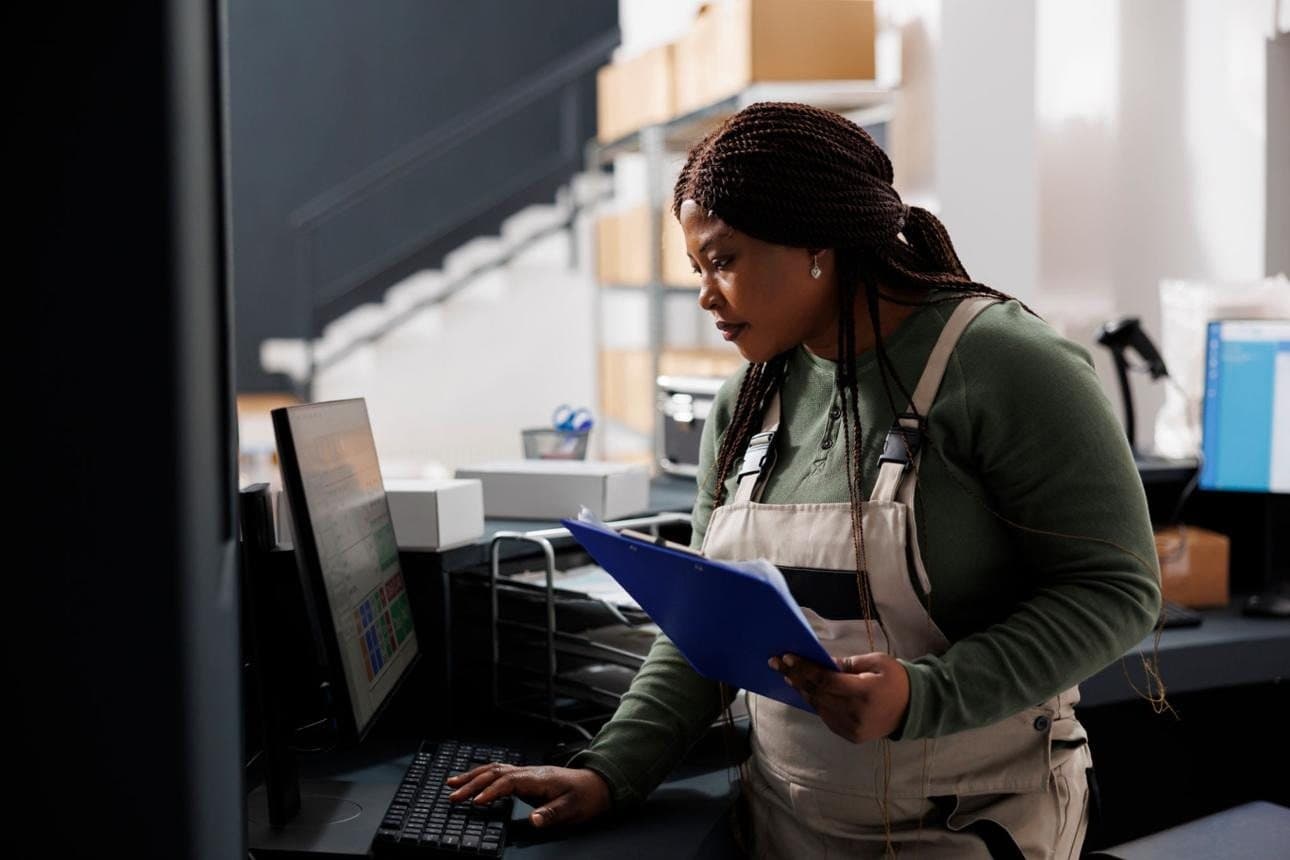 A worker holding a clipboard at a packing workstation, checking a monitor with boxes and shelving behind her.