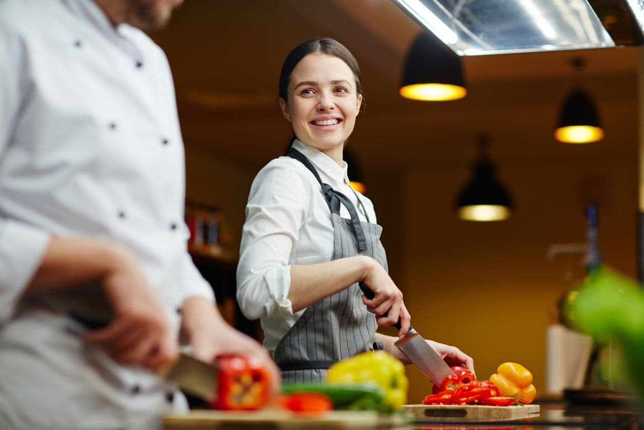 A smiling cook slicing peppers in a warm restaurant kitchen.